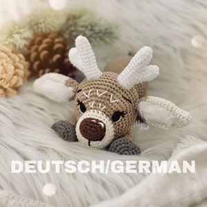 Könnte beinhalten: Ein gehäkeltes Rentier-Spielzeug mit braunem und weißem Fell, das auf einer weißen, flauschigen Oberfläche liegt. Das Rentier hat eine braune Nase, schwarze Augen und ein gesticktes Lächeln. Der Text "DEUTSCH/GERMAN" ist am unteren Rand des Bildes sichtbar.