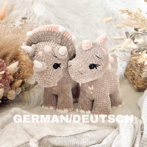 Könnte beinhalten: Zwei gehäkelte Plüsch-Triceratops-Dinosaurier, einer rosa und einer grau, mit gestickten Augen und rosa Akzenten. Die Dinosaurier sitzen auf einer weißen, flauschigen Oberfläche.