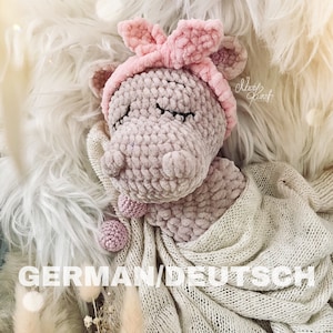 May include: A pink crocheted hippopotamus toy with a pink headband and a white blanket. The toy has closed eyes and a small pink ball in its mouth. The text "Made with Love" is visible in the upper right corner of the image.