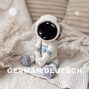 Könnte beinhalten: Handgefertigte Astronautenpuppe aus Häkelarbeit in Weiß, Grau und Schwarz. Die Puppe hat einen detaillierten Astronautenhelm mit schwarzem Visier und grauen Akzenten. Die Puppe sitzt auf einer strukturierten Decke.