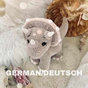 Könnte beinhalten: Ein gehäkeltes graues und rosa Triceratops-Dinosaurierspielzeug mit weißen Akzenten. Das Spielzeug hat eine strukturierte Oberfläche und steht auf einer weißen Oberfläche.