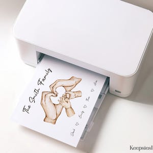 May include: A white printer outputting a sheet of paper featuring a watercolor illustration of adult and child hands forming a heart shape. The text reads The Smith Family, with the names Jack, Emily, Theo, and Ava separated by heart icons.