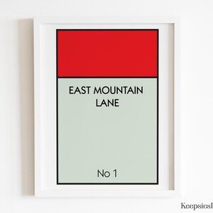 May include: A framed wall art print featuring a rectangular design with a solid red top bar and a light grey lower section. The text EAST MOUNTAIN LANE is printed in black in the centre, with No 1 at the bottom, all enclosed in a white frame.