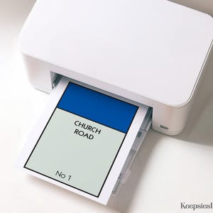 May include: A white desktop printer outputting a rectangular sheet of paper featuring a blue header bar above a light grey field with the black text CHURCH ROAD and the text No 1 at the bottom, all enclosed within a thick black border frame.