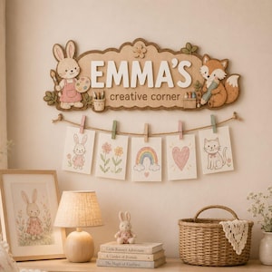 Könnte beinhalten: Ein hölzernes Wandschild mit einem Hasen und einem Fuchs sowie dem Text EMMA'S creative corner. Darunter hält eine Schnur mit fünf Wäscheklammern fünf kleine Zeichnungen eines Hasen, Blumen, eines Regenbogens, eines Herzens und einer Katze.