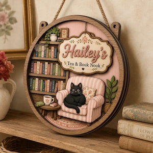 Puede incluir: Un adorno de pared circular de madera con una escena 3D en capas de una habitación acogedora. El letrero dice Hailey's Tea & Book Nook. Incluye una estantería llena de libros coloridos, un sillón de rayas rosas, un gato negro y una planta en maceta.