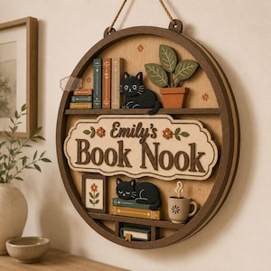 Puede incluir: Un letrero de pared circular de madera con el texto Emily's Book Nook. El diseño incluye dos gatos negros, libros en miniatura, una planta en maceta, un cuadro de flores y una taza de café con vapor, dispuestos en estantes dentro del marco.