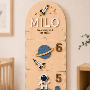 May include: A light wood arched growth chart featuring the name MILO, the text dream beyond the stars, and space-themed decorations including a rocket, a planet with rings, and an astronaut. The chart includes numbered markings for height measurement.