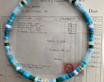 Op de afbeelding: Een blauwe en groene kralenketting met gouden accenten. De ketting ligt op een wit vel papier met tekst die de woorden "Paris le 9 Decembre" en "Doit" bevat.