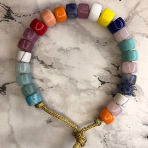 May include: A colorful beaded bracelet with a rainbow of colors, including red, orange, yellow, green, blue, and purple. The bracelet is made with a gold cord and has a knot at the end.