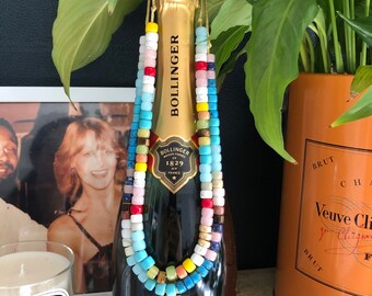 Op de afbeelding: Een fles Bollinger champagne met een kleurrijke kralenketting om de hals. De ketting is gemaakt van turquoise, gele, witte, roze, rode en blauwe kralen.