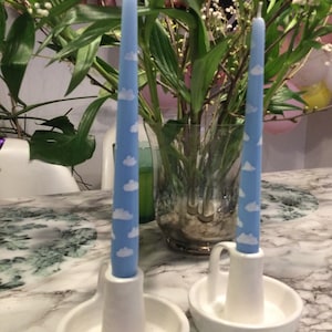 Op de afbeelding: Twee blauwe kaarsen met witte wolkenpatronen staan in witte keramische kandelaars. De kaarsen zijn lang en slank en de houders hebben een klein handvat. De achtergrond omvat groen en een oppervlak met marmerpatroon.