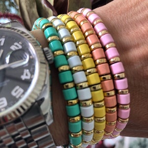 May include: A stack of five colourful bracelets with gold accents. The bracelets are made of small, square beads in shades of pink, orange, yellow, white and green.
