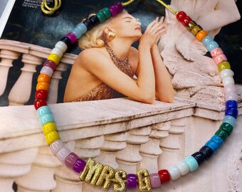 Op de afbeelding: Een kleurrijke kralenketting met een gouden bedel die "MRS G" zegt. De ketting is gemaakt van kleine, ronde kralen in verschillende kleuren, waaronder rood, oranje, geel, groen, blauw en paars. De bedel is van verguld metaal en is met een gouden ketting aan de ketting bevestigd.