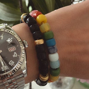 May include: Two bracelets on a wrist. The top bracelet is made of black beads with gold accents. The bottom bracelet is made of colorful beads in shades of pink, yellow, green, blue, and white.