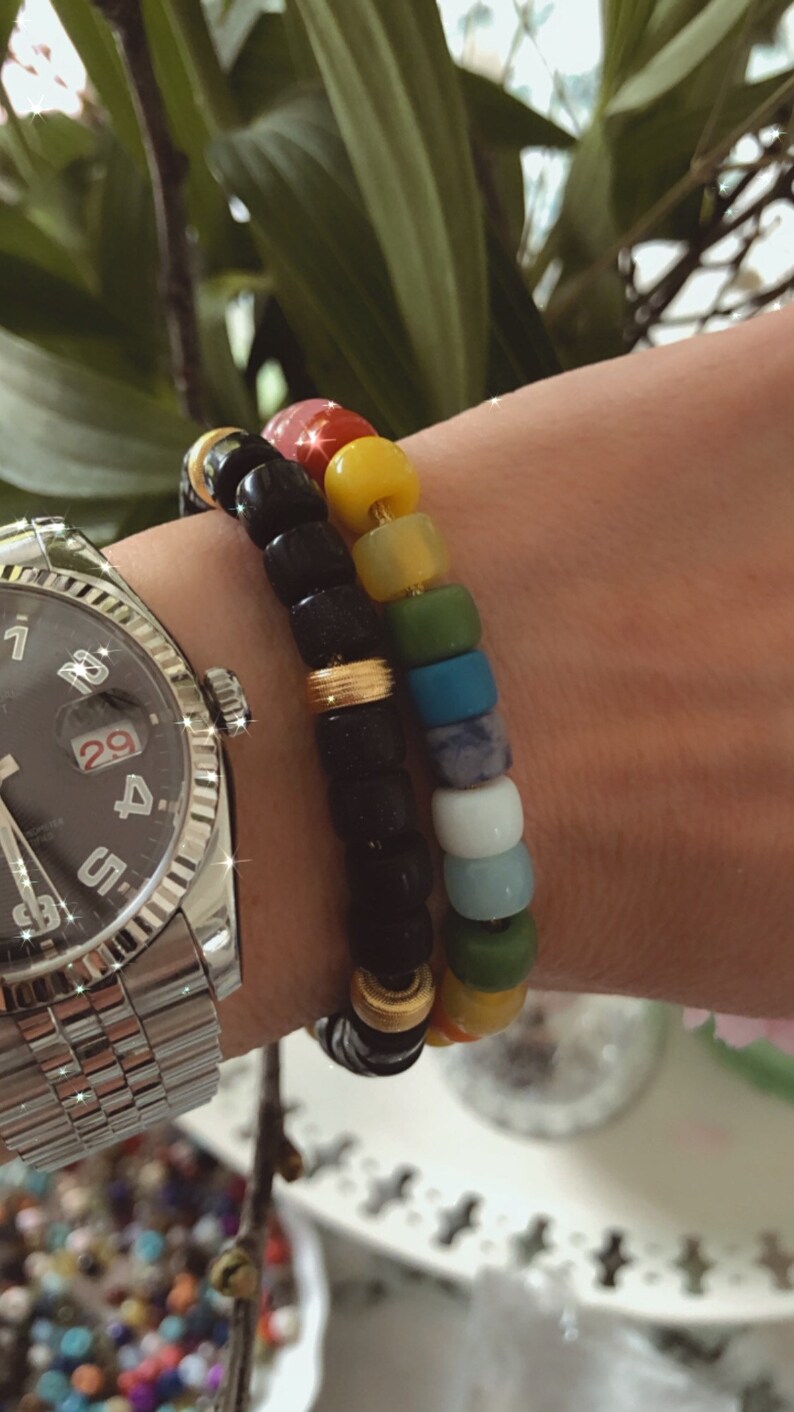 May include: Two bracelets on a wrist. The top bracelet is made of black beads with gold accents. The bottom bracelet is made of colorful beads in shades of pink, yellow, green, blue, and white.