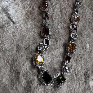 Puede incluir: Un collar de cadena de plata con una variedad de piedras preciosas de colores, incluyendo verde, negro, rojo, amarillo y marrón. Las piedras preciosas están engastadas en plata y tienen un acabado brillante.