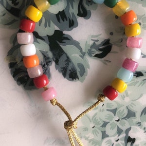 May include: A colorful beaded bracelet with a rainbow of colors, including red, orange, yellow, green, blue, and purple. The bracelet is made with small, round beads and has a gold cord with a knot at the end.