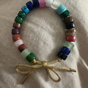 May include: A colorful beaded bracelet with a gold cord tie. The beads are made of various stones, including turquoise, malachite, and amethyst.