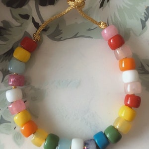 May include: A colorful beaded bracelet with a rainbow of colors, including pink, red, orange, yellow, green, blue, and white. The bracelet is made with round beads and has a gold cord.