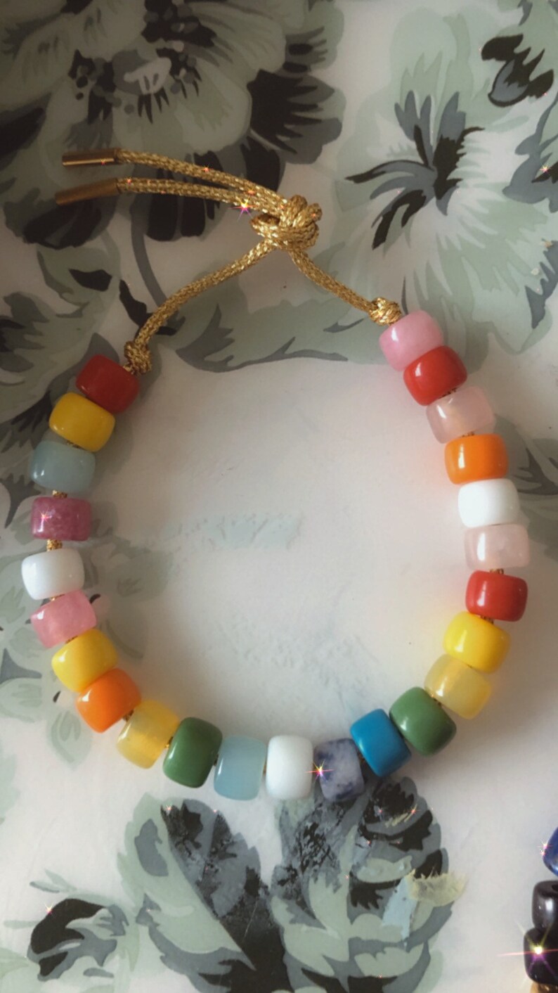 May include: A colorful beaded bracelet with a rainbow of colors, including pink, red, orange, yellow, green, blue, and white. The bracelet is made with round beads and has a gold cord.