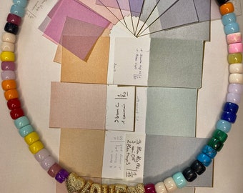 Op de afbeelding: Een kleurrijke kralenketting met een gouden "LOVE"-bedel. De kralen zijn in een regenboogpatroon en de ketting ligt op een kleurenkaart.