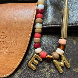 May include: A beaded keychain with a gold-coloured clasp and charms. The beads are in shades of brown, red, white, and pink. Gold-coloured letters spell out "EM". The keychain is attached to a brown leather item.