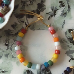 May include: A colorful beaded bracelet with a rainbow of colors, including red, orange, yellow, green, blue, and purple. The bracelet is made with small, round beads and has a gold cord closure.