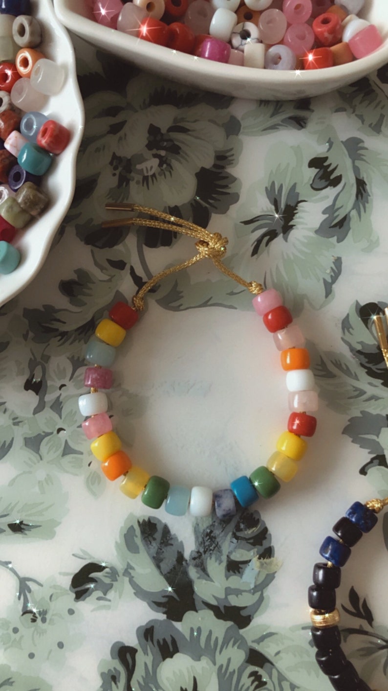 May include: A colorful beaded bracelet with a rainbow of colors, including red, orange, yellow, green, blue, and purple. The bracelet is made with small, round beads and has a gold cord closure.