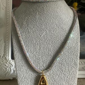 Puede incluir: Un conjunto de collar y gargantilla de brillantes. El collar presenta un colgante dorado con la forma de la letra "A". Las joyas se exhiben sobre una cabeza de maniquí gris.