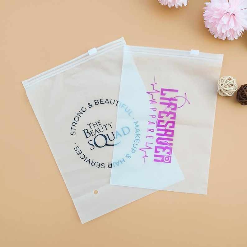 May include: Two clear plastic bags with zipper closures. One bag has the text "The Beauty Squad" in a circle with the text "Strong & Beautiful. Hair & Makeup Services."  The other bag has the text "Lifesaver" in pink with a heart beat symbol and the text "Apparel, Makeup & Hair".