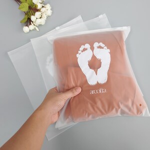 Puede incluir: Bolsa de pl&aacute;stico transparente con cremallera, dise&ntilde;o de huellas blancas y el texto "arcofta".