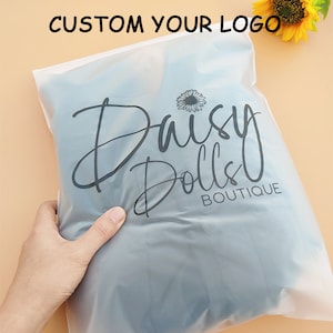 Puede incluir: Una bolsa de pl&aacute;stico transparente con un logotipo en blanco y negro que dice "Daisy Dolls Boutique".