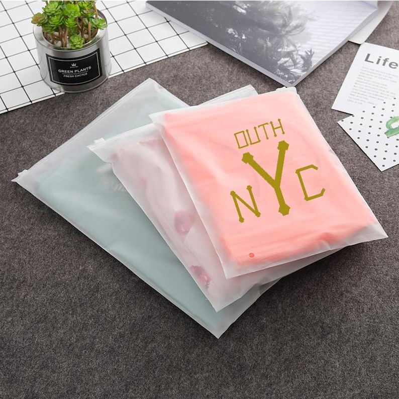 May include: Three clear plastic zippered bags with a pink shirt inside the top bag. The shirt has the text "OUTH NYC" printed in gold.