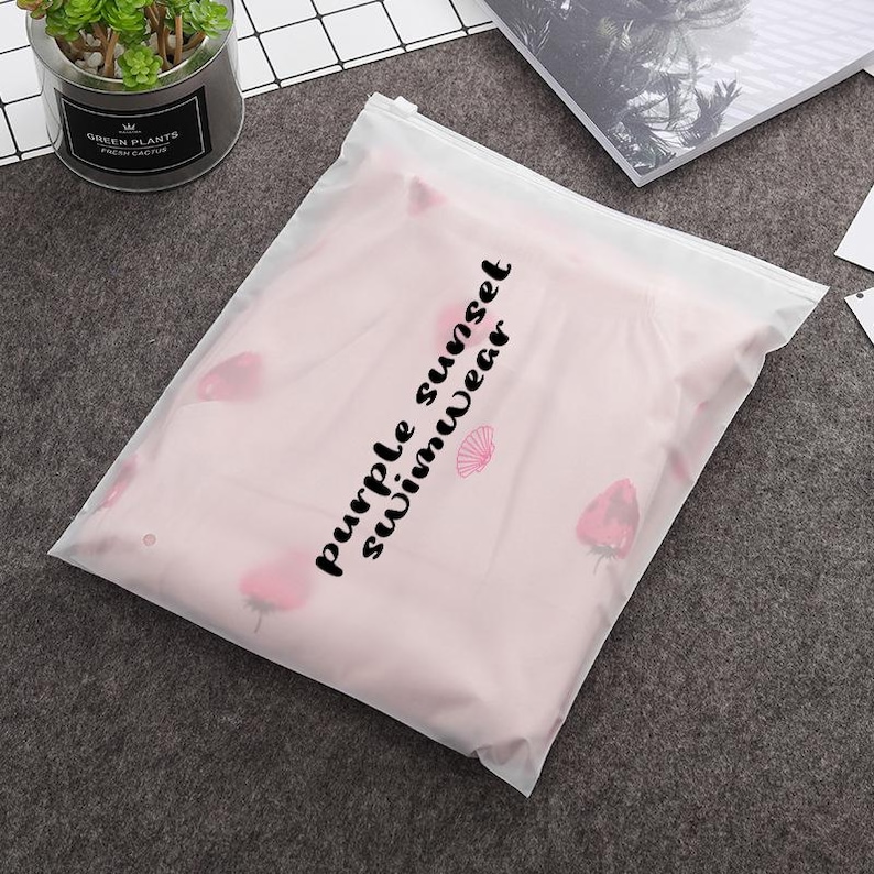 May include: A clear plastic bag with a zipper closure. The bag contains a pink swimsuit with a floral print and the text "purple sunset swimwear" printed on the front.