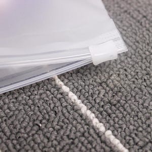 May include: A clear plastic zippered bag with a white slider. The bag is lying on a grey and white textured surface.