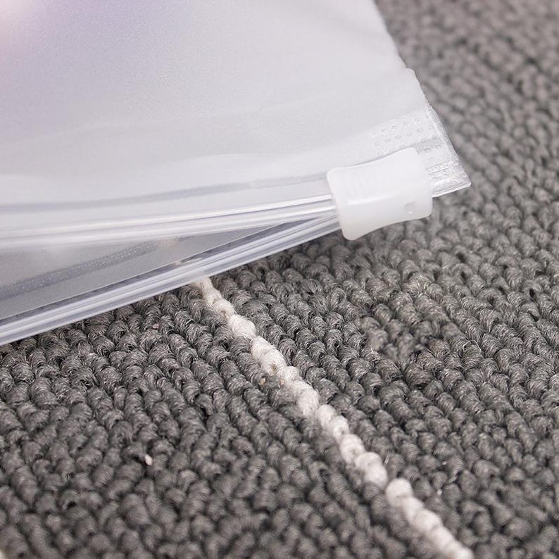 May include: A clear plastic zippered bag with a white slider. The bag is lying on a grey and white textured surface.
