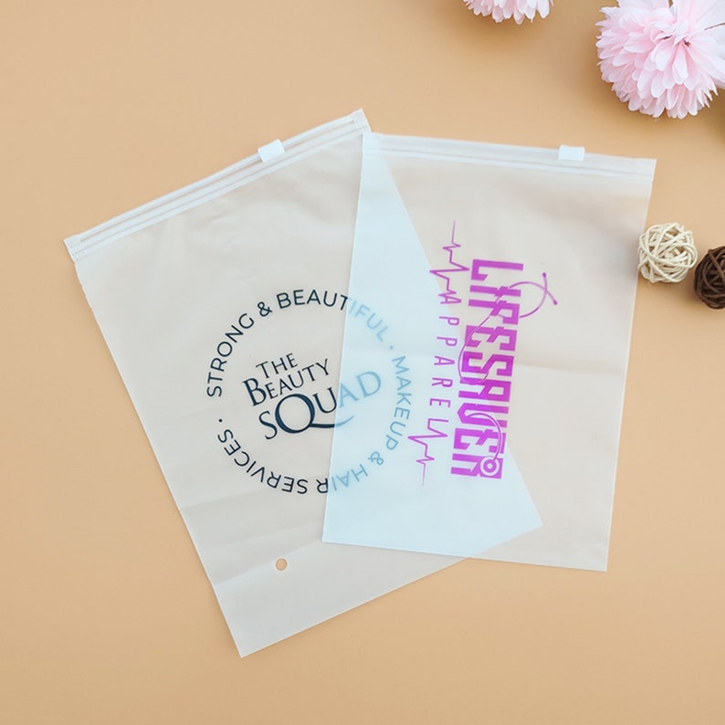 Puede incluir: Dos bolsas de pl&aacute;stico transparentes con cremallera y diferentes logotipos impresos en ellas. La bolsa de la izquierda tiene un logotipo circular negro con el texto "The Beauty Squad" y el texto "Strong & Beautiful. Hair & Makeup Services". La bolsa de la derecha tiene un logotipo rosa con el texto "Lifesaver" y el texto "Hair Apparel Makeup".