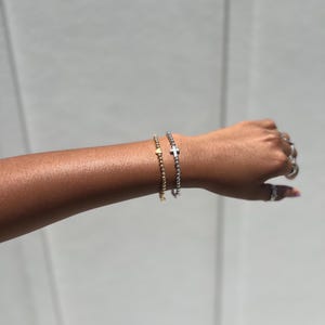 May include: Two beaded bracelets on a person's wrist. One bracelet has a gold heart charm and the other has a silver cross charm.