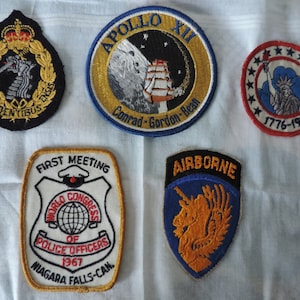 May include: Five embroidered patches, including a black and gold patch with a crown and a dragon, a gold and blue patch with the text "APOLLO XII Conrad-Gordon-Bean", a red, white and blue patch with a star-shaped border and the Statue of Liberty, a white patch with a gold border and the text "FIRST MEETING WORLD CONGRESS OF POLICE OFFICERS 1967 NIAGARA FALLS-CAN.", and a blue and gold patch with the text "AIRBORNE" and a unicorn.