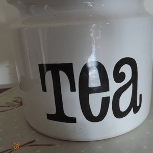 May include: White ceramic tea jar with a cork lid and black lettering that says "Tea".