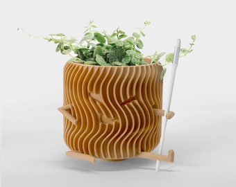 May include: A light brown, cylindrical planter with a wavy, vertical ribbed texture and a carved face. The planter features small, attached arms and legs, with one arm holding a thin, white stylus. A green, trailing plant grows from the top.