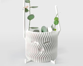 May include: A white cylindrical planter with a vertical wavy texture and two integrated tall support rods. A green vining plant with small round leaves grows upward, secured to one of the rods with a thin piece of twine. The planter stands on three small feet.