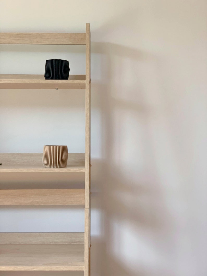 May include: A wooden ladder shelf with two shelves, one with a black planter and the other with a light brown planter.