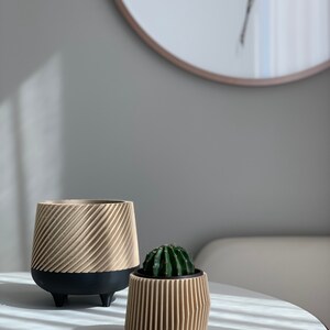 May include: Two beige ceramic planters with a ribbed design. The larger planter has black legs and a small cactus plant is in the smaller planter.