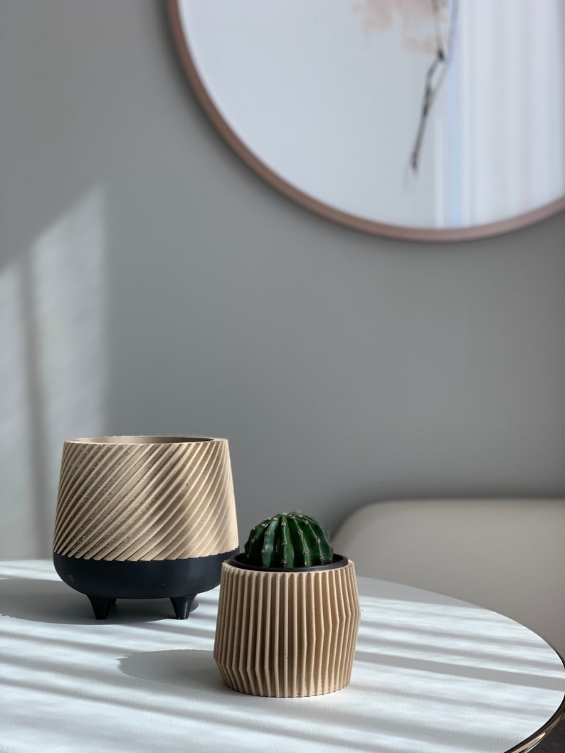 May include: Two beige ceramic planters with a ribbed design. The larger planter has black legs and a small cactus plant is in the smaller planter.