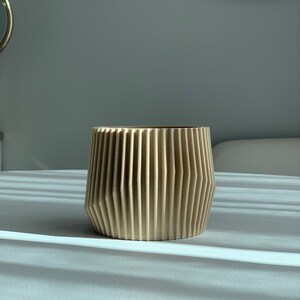 May include: A light brown, wooden planter with a geometric, ribbed design. The planter is empty and sitting on a white surface.