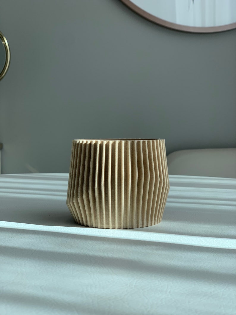 May include: A light brown, wooden planter with a geometric, ribbed design. The planter is empty and sitting on a white surface.
