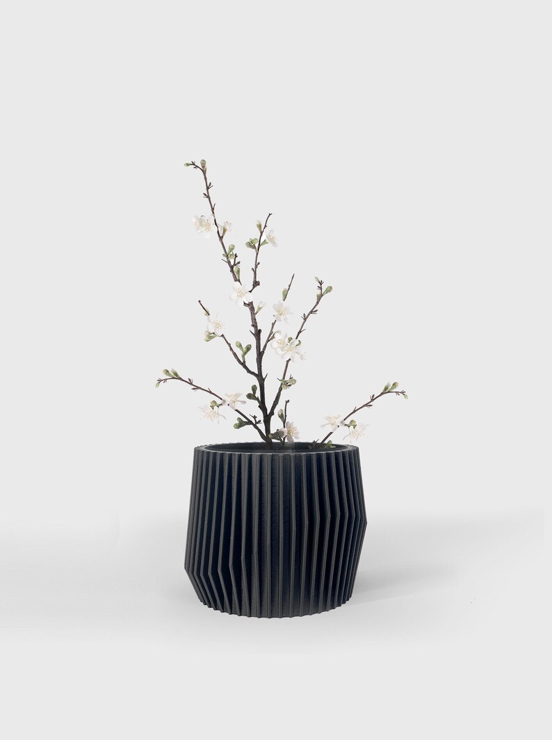 May include: A black, ribbed ceramic planter with a small branch of white blossoms.