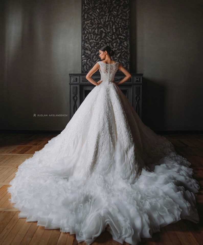 May include: A white wedding dress with a long train and a lace bodice. The dress is fitted at the waist and flows out into a full skirt. The train is made of layers of tulle and has a ruffled edge.